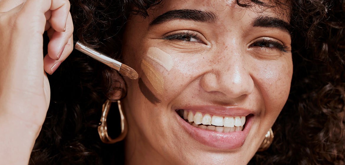 Das Gesicht einer lachenden Frau mit dunklen Locken und schweren goldenen Ohrringen von vorne. Sie trägt mit ihrer rechten Hand mittels eines Makeup-Bürstchens drei breite Striche Makeup auf ihre rechte Wange auf, wobei der oberste sehr hell und der unterste sehr dunkel ist.